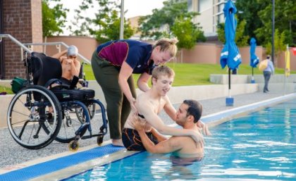 A young man is being helped from a wheelchair into a swimming pool by two trainers.